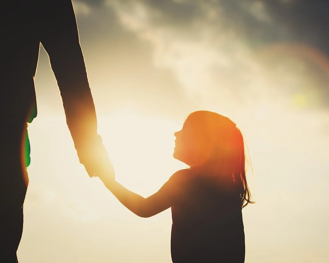 A child holding an adult's hand, silhouetted against warm sunlight